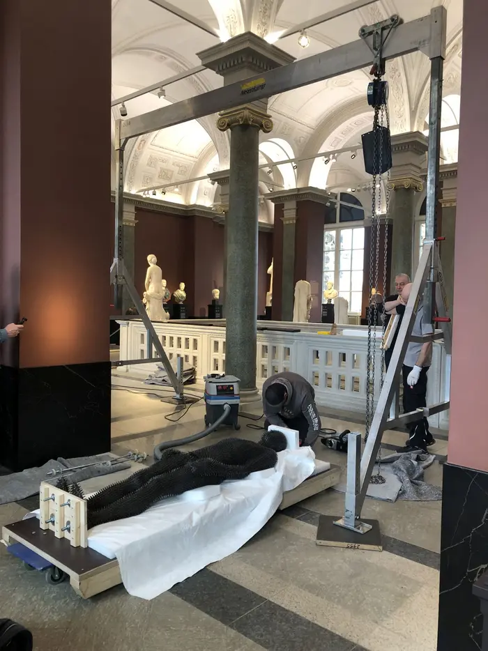 Zu sehen ist das Werk "Under my Skin" von Antony Gormley aus dem Jahr 1997, einem in Eisen ausgeführten einstigen Abgusses des Künstlers. Es ist gänzlich von stacheligen Eisenstiften überzogen. Auf dem Foto wird es liegend vorbereitet um in der antikenhalle installiert zu werden.