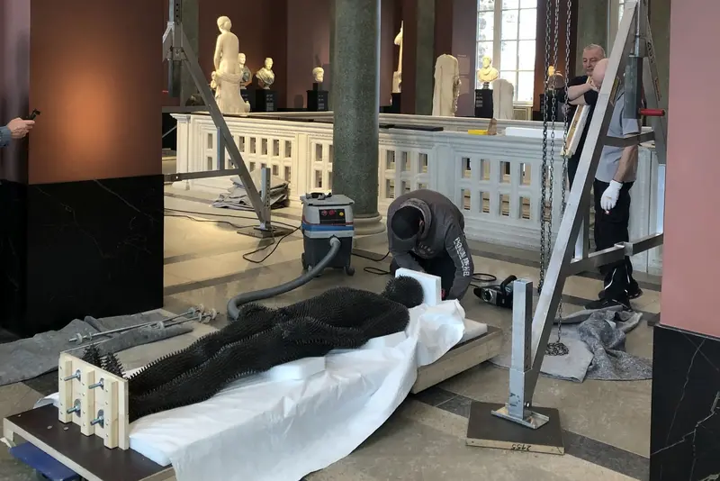 Zu sehen ist das Werk "Under my Skin" von Antony Gormley aus dem Jahr 1997, einem in Eisen ausgeführten einstigen Abgusses des Künstlers. Es ist gänzlich von stacheligen Eisenstiften überzogen. Auf dem Foto wird es liegend vorbereitet um in der antikenhalle installiert zu werden.