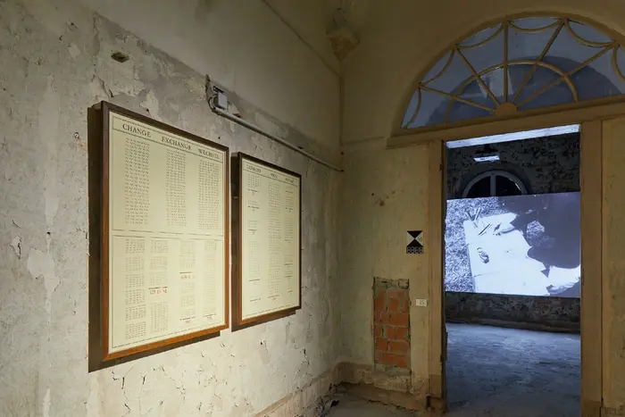 Zu sehen ist ein zweiteiliges Werk von Marcel Broodthaers, das an der Wand hängt, durch die Tür ist das Video "La Pluie" vom selben Künstler zu sehen.