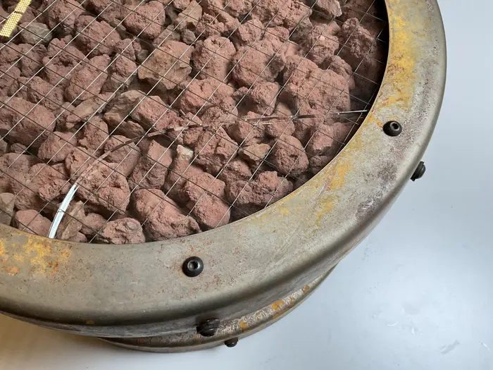 Die Wandinstallation "Catalog: Terra Firma Nineteen Hundred Eighty Nine, #3" aus dem Jahr 1989 von Ashley Bickerton besteht aus zwölf symmetrisch gehangen Bullaugen, die mit unterschiedlichsten Materialien wie Erdnussflips, Zigarettenstummel, Flechten und Korallen gefüllt sind. Auf dem Foto ist ein Detail des mit Steinen gefüllten Behältnisses zu sehen, welches einen Sprung im Glas hat. 