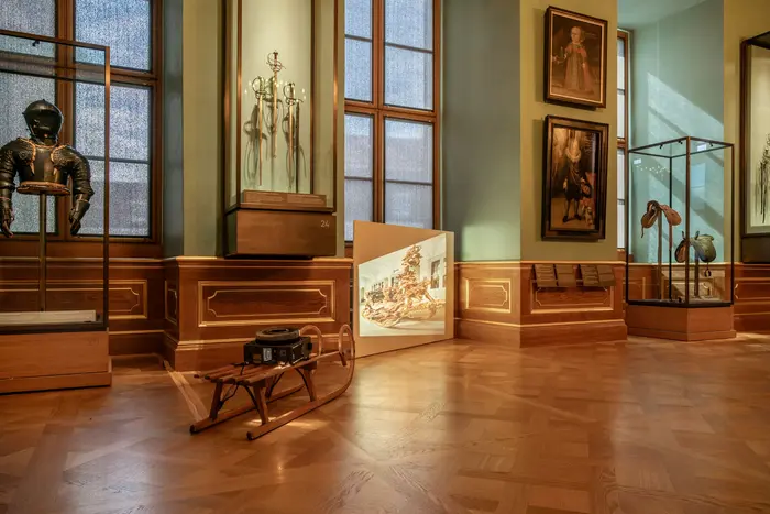 In einem der Paradeappartements steht ein Diaprojektor auf einem herkömmlichen hölzernen Schlitten. Es wird ein Abbild des zeremoniellen Prunkschlittens von König Ludwig II. von Bayern in verzerrter Weise an die Wand projiziert. 