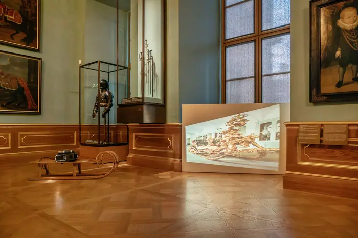 In einem der Paradeappartements steht ein Diaprojektor auf einem herkömmlichen hölzernen Schlitten. Es wird ein Abbild des zeremoniellen Prunkschlittens von König Ludwig II. von Bayern in verzerrter Weise an die Wand projiziert. 