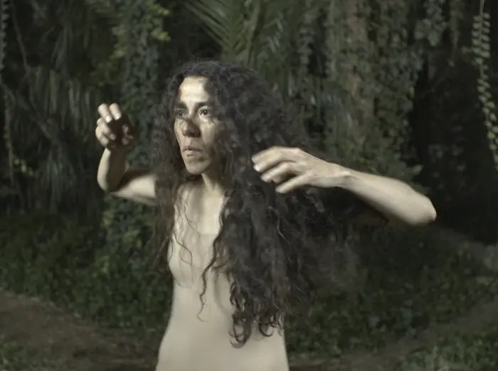 Eine Frau mit dunklem lockigen Haar macht Übungsbewegungen vor dem Hintergund eines botanischen Gartens.