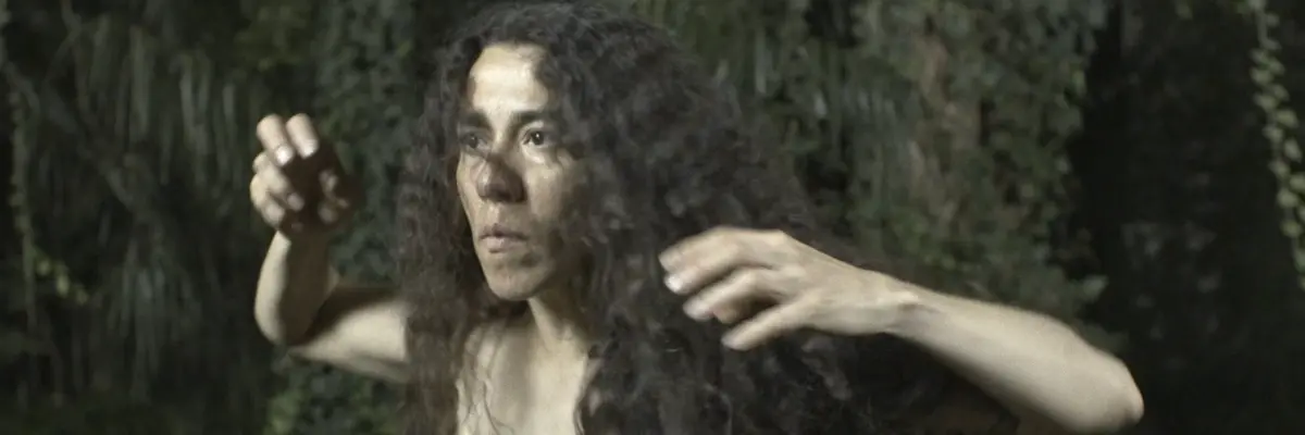 Eine Frau mit dunklem lockigen Haar macht Übungsbewegungen vor dem Hintergund eines botanischen Gartens.