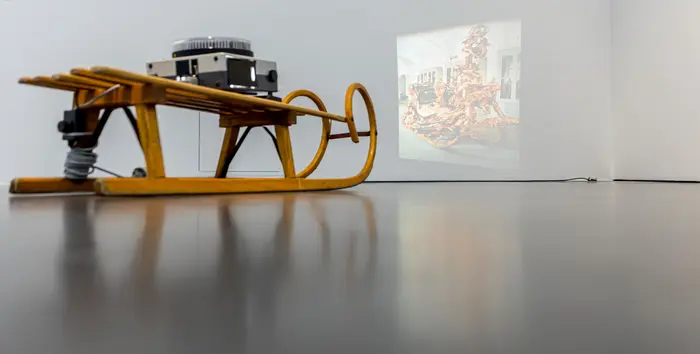 Im Ausstellungsraum steht ein Diaprojektor auf einem herkömmlichen hölzernen Schlitten. Es wird ein Abbild des zeremoniellen Prunkschlittens von König Ludwig II. von Bayern in verzerrter Weise an die Wand projiziert. 