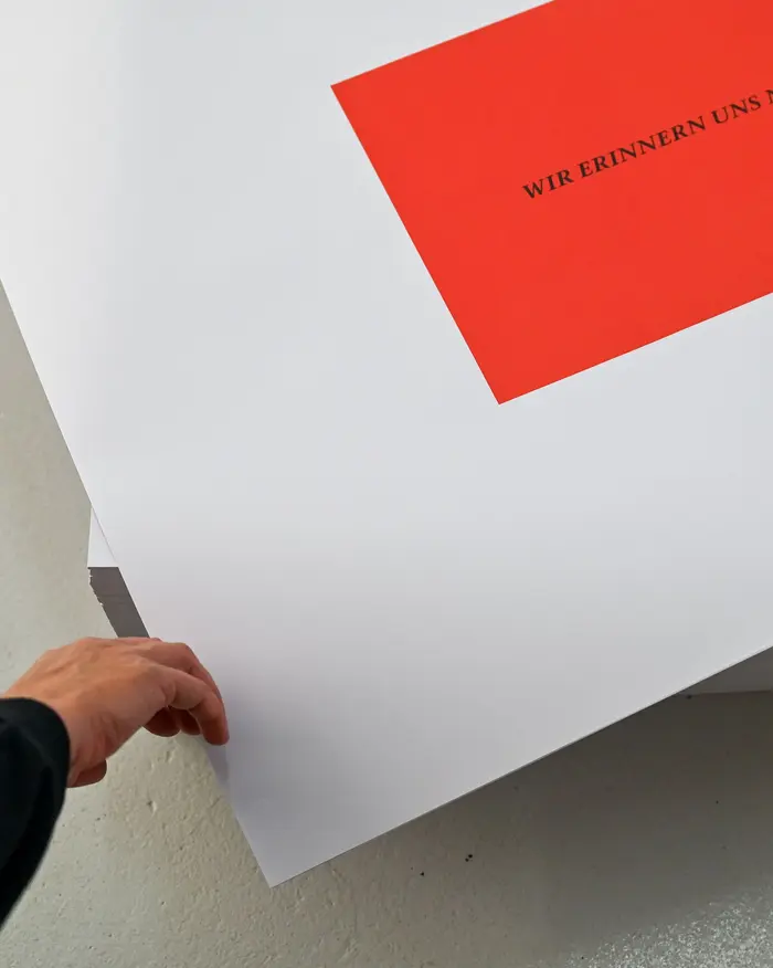 Eine Hand greift nach einem Poster eines Stapels. Auf dem Blatt steht auf rotem Grund der Satz "Wir erinnern uns nicht". Das Wekr gehört zu einer wichtigen Riehe des Künstlers Felix Gonzalez-Torres.