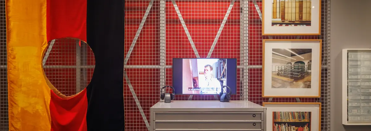 Ausstellungsansicht der Kunstkammer Gegenwart im Residenzschloss Dresden im Dezember 2023. Die Ausstellungsarchitektur erinnert an ein Depot und ist modular aufgebaut.  Zu sehen ist ein Videobildschirm mit "Rückblick" von Janet Grau, ein Grafikschrank mit Werken von Francisco de Goya und den Chapman Brüdern, eine DDR-Flagge, aus der mittig ein Loch ausgeschnitten wurde mit dem Titel „Weil Sie zwischen den Beinen anders aussehen (German Problem – Not Yours)“ von Georg Herold, sowie eine drei Fotografien von Cadida Höfer in den Räumen der Staatlichen Kunstsammlungen Dresden im Jahr 2002.