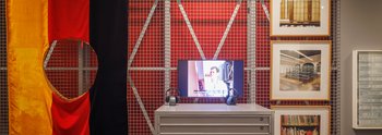 Ausstellungsansicht der Kunstkammer Gegenwart im Residenzschloss Dresden im Dezember 2023. Die Ausstellungsarchitektur erinnert an ein Depot und ist modular aufgebaut.  Zu sehen ist ein Videobildschirm mit &quot;Rückblick&quot; von Janet Grau, ein Grafikschrank mit Werken von Francisco de Goya und den Chapman Brüdern, eine DDR-Flagge, aus der mittig ein Loch ausgeschnitten wurde mit dem Titel „Weil Sie zwischen den Beinen anders aussehen (German Problem – Not Yours)“ von Georg Herold, sowie eine drei Fotografien von Cadida Höfer in den Räumen der Staatlichen Kunstsammlungen Dresden im Jahr 2002.