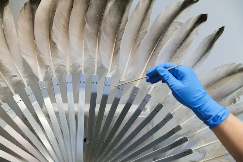 Zu sehen ist die Hand der Restauratorin beim Entstauben einer Feder an dem Werk "Silver Crane" von Rebecca Horn aus dem Jahr 1984. Das kinetische Werk besteht aus einem Federkranz aus Kranichfedern, die von einem Motor betrieben sich öffnen und schließen.
