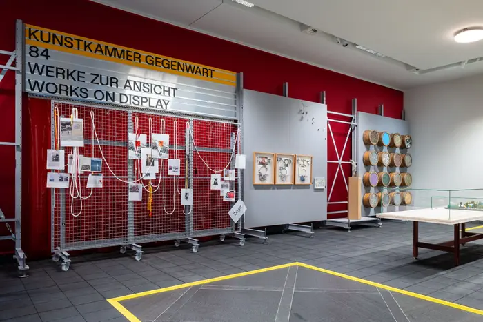 Das Bild zeigt eine an ein Depot angelehnte Ausstellungsarchitektur mit Werken der Schenkung Sammlung Hoffmann, des Kunstfonds, des Kupferstich-Kabinetts und des Albertinums. Die Ausstellungsansicht zeigt Wanderblog von Eran Schaerf, Scherenschnitte von Felix Droese, „Statement“ von Georg Herold, das Erste Modell für die Kunsthalle Dresden, sowie ein Entwurf auf Papier von Frank Stella, und „Catalog: Terra Firma Nineteen Hundred Eighty Nine, #3“ von Ashley Bickerton.