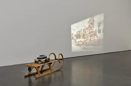 Im Ausstellungsraum steht ein Diaprojektor auf einem herkömmlichen hölzernen Schlitten. Es wird ein Abbild des zeremoniellen Prunkschlittens von König Ludwig II. von Bayern in verzerrter Weise an die Wand projiziert. 