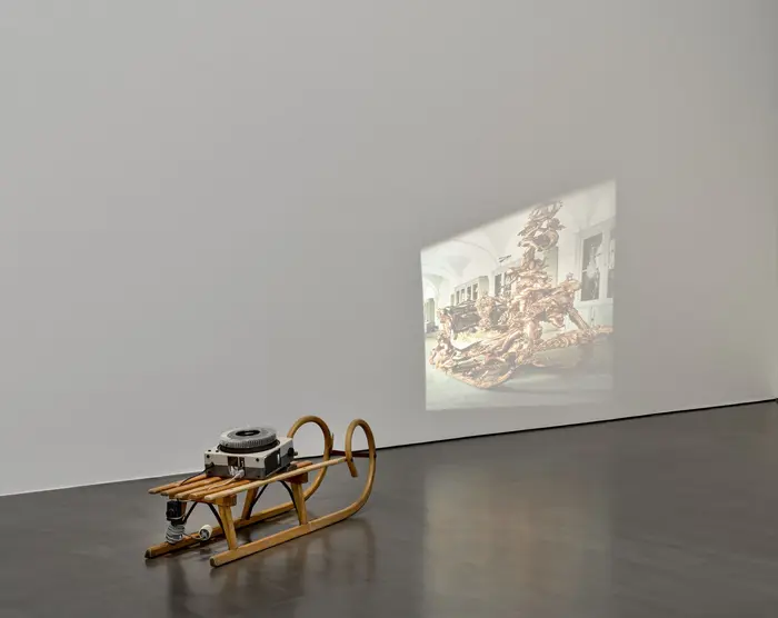 Im Ausstellungsraum steht ein Diaprojektor auf einem herkömmlichen hölzernen Schlitten. Es wird ein Abbild des zeremoniellen Prunkschlittens von König Ludwig II. von Bayern in verzerrter Weise an die Wand projiziert. 