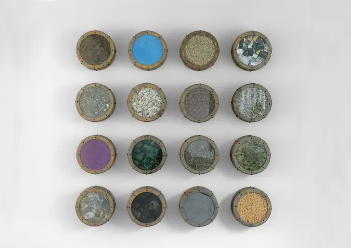 Die Wandinstallation "Catalog: Terra Firma Nineteen Hundred Eighty Nine, #3" aus dem Jahr 1989 von Ashley Bickerton besteht aus zwölf symmetrisch gehangen Bullaugen, die mit unterschiedlichsten Materialien wie Erdnussflips, Zigarettenstummel, Flechten und Korallen gefüllt sind. 