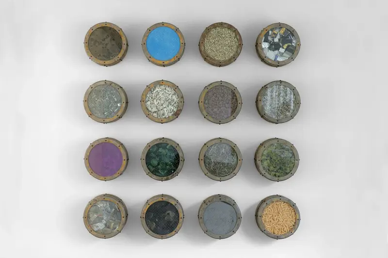 Die Wandinstallation "Catalog: Terra Firma Nineteen Hundred Eighty Nine, #3" aus dem Jahr 1989 von Ashley Bickerton besteht aus zwölf symmetrisch gehangen Bullaugen, die mit unterschiedlichsten Materialien wie Erdnussflips, Zigarettenstummel, Flechten und Korallen gefüllt sind. 