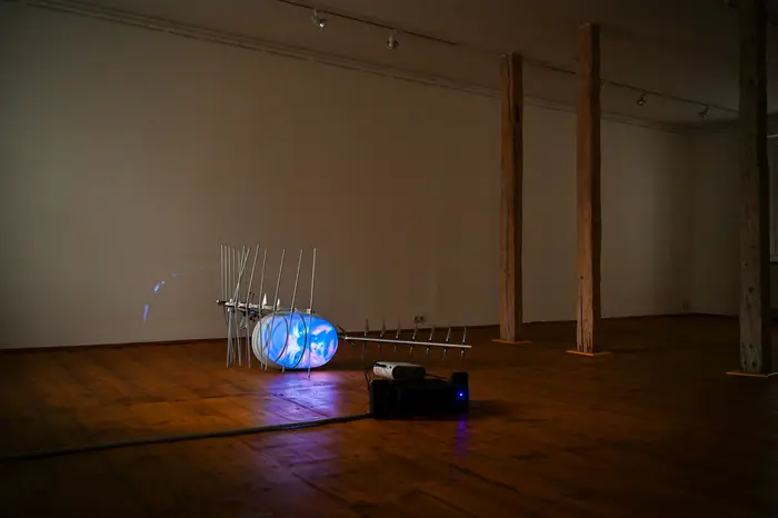 Eine Videoskulptur des Künstlers Tony Oursler bestehend aus einem metallenen Empfänger und einer auf dem Boden liegenden Projektionsfläche in Form eines Kopfes, der einen theatralen Monolog hält.