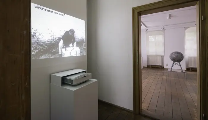 Zu sehen ist eine Videoarbeit des Künstlers Marcel Broodthaers mit dem Titel "La Pluie (der Regen". Hier versucht der Künstler im Garten sitzend etwas mit Tinte aufzuschreiben, während es in Strömen auf ihn herabregnet, was den Schaffensprozess erschwert.