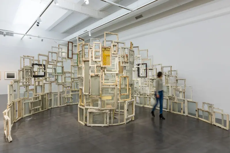 Zu sehen ist das Werk "Inside-Outside" der Künstlerin Chiharu Shiota aus dem Jahr 2009. Die begehbare Installation besteht aus Altbaufenstern, die die Künstlerin in Ost-Berlin zusammengetragen und meterhoch spiralförmig angeordnet hat. In der Mitte steht ein Stuhl auf dem man von innen nach außen schauen kann.