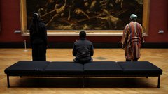 Drei schwarze Männer sitzen und stehen in einigem Abstand auf einer Bank im Museum und betrachten ein großformatiges Historiengemälde