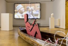 Ausstellungsraum art gluchowe e.V. mit roter bemalter Holzfigur in einem Holzboot, im hintergrund Projektion eines Videos von Marcel Odenbach